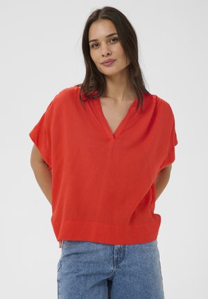 Hosszú sötét hajú nő, laza, piros V-nyakú inget és kék farmert visel, egyszínű világos háttér előtt áll.