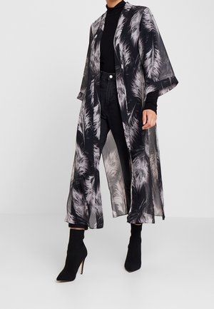 Personne portant un long manteau transparent noir et blanc à motif plume sur un col roulé noir, un jean noir et des bottines noires à talons hauts.