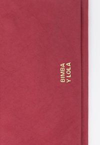 Bolso textil rojo con una textura suave, que presenta un logo dorado de "BIMBA Y LOLA" en el lateral. Diseño simple y rectangular.