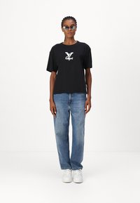 Zwarte katoenen oversized t-shirt met een wit logo op de borst, gecombineerd met lichtblauwe denim jeans en witte sneakers. Strakke zonnebril inbegrepen.