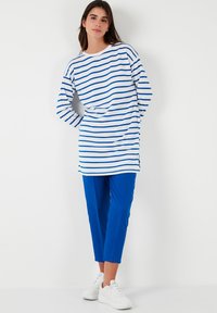 Túnica de manga larga, a rayas blancas y azules, combinada con pantalones azules y zapatillas blancas. La túnica tiene un escote redondo y aberturas laterales.