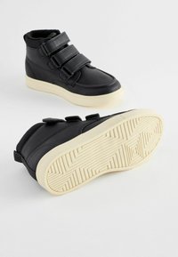Baskets montantes noires avec trois bandes Velcro, tige en cuir lisse, semelle en caoutchouc avec motif texturé, et semelle de couleur crème.