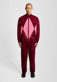 Vīnogu krāsas trikotāžas sporta tērps ar rozā dimanta rakstiem, jaka ar rāvējslēdzēju un augstu apkakli, kā arī atbilstošas bikses ar izsmalcinātām līniju detaļām.