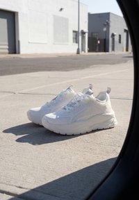 Paire de baskets blanches posées sur un trottoir en béton à l'extérieur près de bâtiments industriels en plein jour.