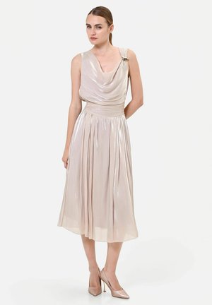 Donna con coda di cavallo che indossa un abito midi beige lucido senza maniche con scollatura drappeggiata e tacchi alti abbinati su sfondo neutro.
