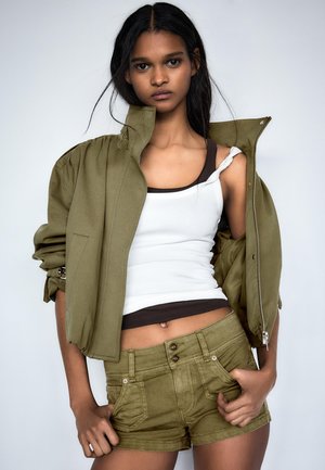 Mladá žena má na sebe olivovo zelenú bundu spadnutú z ramien, biele tielko a k tomu olivovo zelené šortky, stojí pred jednofarebným pozadím.