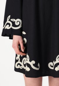 Μαύρο φόρεμα με λευκά κεντημένα κυκλικά σχέδια στο τελείωμα και στα μανίκια. Το ύφασμα φαίνεται απαλό με χαλαρή εφαρμογή και φαρδιά σχεδίαση.