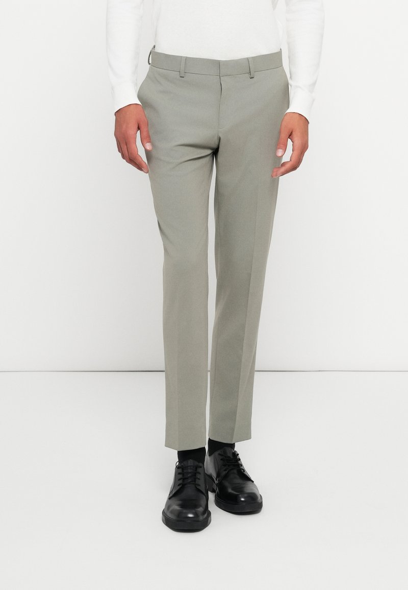 Világosszürke, slim fit szabású nadrág, sima tapintással és övhurkokkal. Fekete fűzős cipővel viselve.