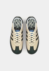 Beige sneakers met zwarte strepen, groene accenten en een gestructureerde rubberen zool. Gevoerde binnenzolen hebben een herhalend logo-patroon.