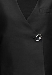 Ένδυμα από μαύρο ύφασμα με σχέδιο V λαιμόκοψης, που διαθέτει ασημένιο οβάλ κούμπωμα και μια μικρή τσέπη στην αριστερή πλευρά. Λεία υφή.