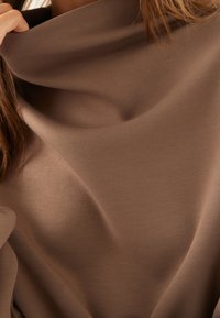 Brązowa bluzka z dekoltem w kształcie cowl, wykonana z miękkiego, elastycznego materiału. Charakteryzuje się gładką teksturą oraz luźnym krojem z delikatnymi fałdami.