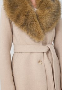 Beeži villane mantel, millel on suur, pehme pruun sääris, vöökoht ja kaks küljetaskut, millel on peen nuppude sulgemine.