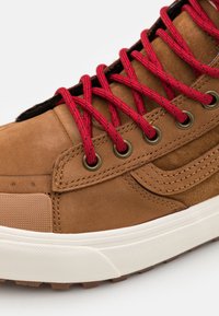Sapatilhas de couro castanho com atacadores vermelhos, ilhós de latão e sola de borracha texturizada. O design inclui uma faixa lateral e detalhes cosidos.