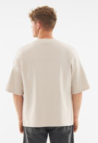 Tricou bej cu mânecă scurtă, fabricat dintr-un material moale, cu o croială lejeră și guler rotund, având o textură netedă și fără modele vizibile.
