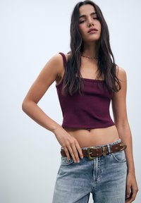 Jonge vrouw met lang, donker haar, gekleed in een wijnrood crop top, lichtblauwe jeans en een bruine leren riem met metalen details.