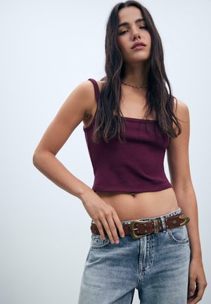 Giovane donna con lunghi capelli scuri che indossa un crop top bordeaux, jeans azzurri chiari e una cintura di pelle marrone con dettagli metallici.