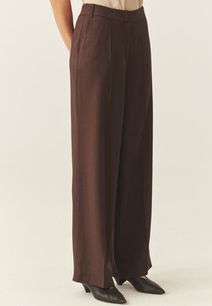 Frau trägt hoch taillierte, weit geschnittene braune Hose kombiniert mit schwarzen spitzen Absätzen, steht vor einem einfarbigen Hintergrund.