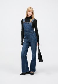 Τζιν σαλοπέτα με χαλαρή εφαρμογή, που διαθέτει μπροστινή τσέπη, ρυθμιζόμενους ιμάντες και φαρδιά πόδια. Φοριέται πάνω από ένα μαύρο μπλούζα με μακριά μανίκια και συνδυάζεται με μαύρη τσάντα.