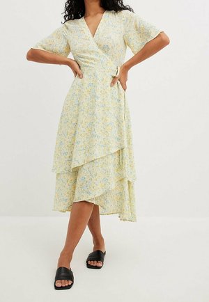 Vestido cruzado floral amarillo con mangas cortas, falda en capas y textura suave. Presenta patrones azules y amarillos, combinado con sandalias negras de tipo slide.