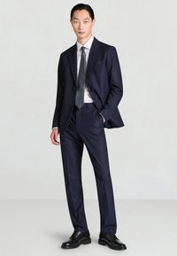 Marineblauer Anzug mit schlankem Schnitt, Stegkragen und einem einzelnen Knopf. Kombiniert mit einem hellen Hemd und gemusterter Krawatte, ergänzt durch schwarze Schuhe.