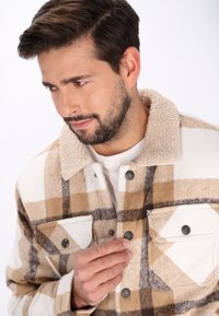 Buffalo plaid takki beige- ja ruskeasävyissä, jossa on sherpa-kaulus, napitettu etuosa ja kaksi läpällistä rintataskua. Pehmeä rakenne.
