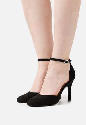 Jambes de femme portant des escarpins en suède noir avec des brides à la cheville et des boucles argentées, sur un fond blanc uni.