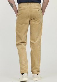 Pantalons en velours côtelé beiges avec une coupe droite, dotés de deux poches arrière et d'une texture côtelée subtile. Portés avec des baskets blanches.
