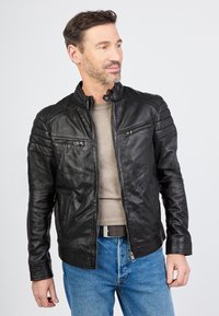 Schwarze Lederjacke mit einem Reißverschluss vorne, Druckknopfkragen, gerippten Schulterdetails und zwei Brusttaschen, getragen über einem grauen Hemd und blauen Jeans.
