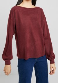 Maglione in borgogna a maglia con scollatura a barca ampia, spalle scese e maniche lunghe e voluminose. Tessuto morbido e dettaglio a coste sull'orlo.