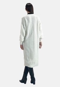 Knielanger weißer Strickcardigan mit gerippter Textur, überschnittenen Schultern und langen Ärmeln. Getragen über engen schwarzen Hosen und Stiefeln.