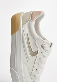 Sneaker bianco con accenti testurizzati sottili in beige e rosa, con lacci bianchi e suola in gomma, esposto in un angolo.