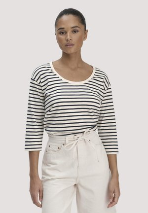 Vrouw draagt een zwart-wit gestreept shirt met driekwart mouwen, ingestopt in een witte broek met hoge taille en een ceintuur, staand tegen een effen achtergrond.