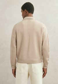 Bēšas krāsas adīts džemperis ar apkakli, ribberiem aprocēm un apakšu, ar teksturētu audumu un smalku vertikālu šuvju detaļu.