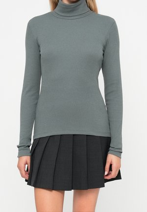 Femme portant un haut à col roulé gris à nervures et une jupe plissée foncée, debout devant un fond blanc uni.