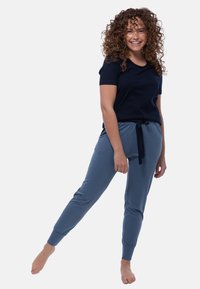 Leende kvinna med lockigt hår som har på sig en marinblå t-shirt och blå joggingbyxor, stående barfota med en ben utsträckt åt sidan.