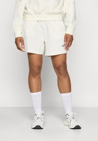 Heledad beez šortsid, mis on valmistatud pehmest kangast ja millel on must tikandiga logo. Kanda valgete sokkide ja heledate tennisetossudega.