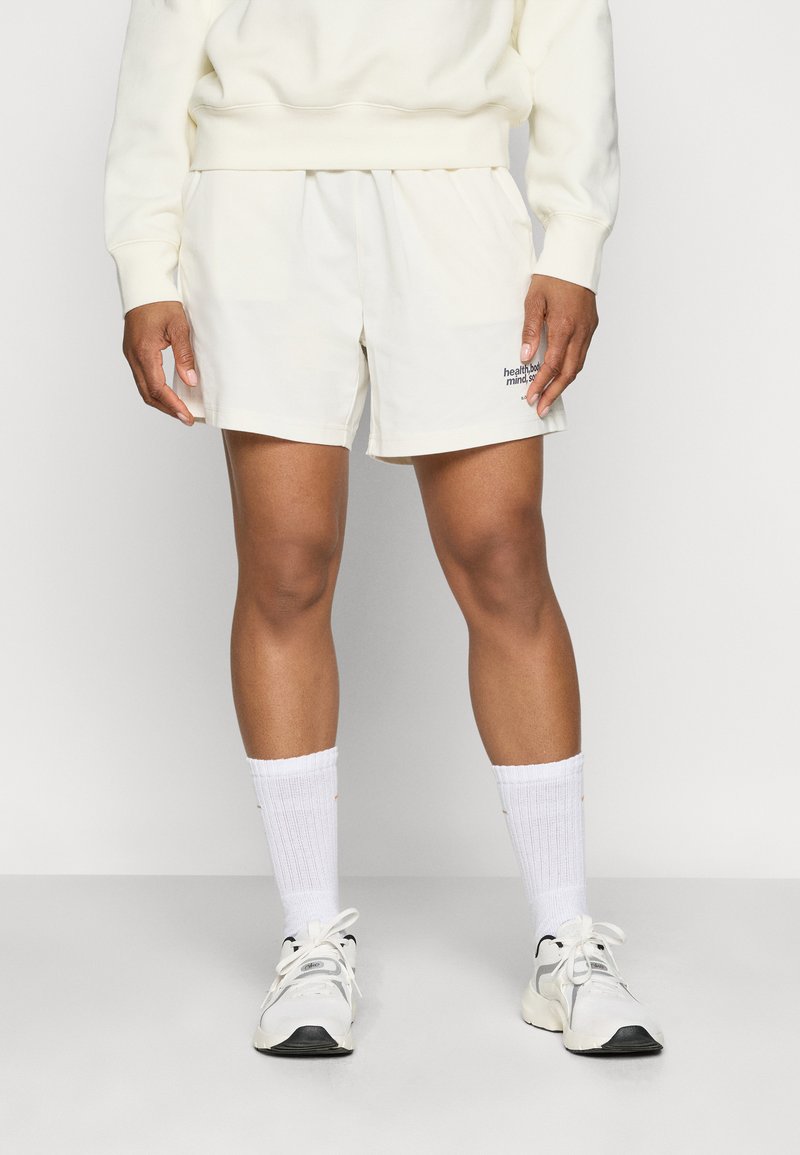 Heledad beez šortsid, mis on valmistatud pehmest kangast ja millel on must tikandiga logo. Kanda valgete sokkide ja heledate tennisetossudega.