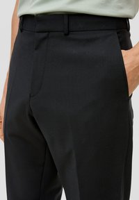 Schwarze Tailored Hose mit glatter Textur, schlanker Passform, Gürtelschlaufen und Seitentaschen, teilweise in der Taille sichtbar.