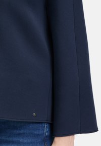 Marineblaues Langarmoberteil aus strukturiertem Stoff, mit seitlichen Schlitzen und einem metallischen Akzent am Saum, kombiniert mit Jeans.