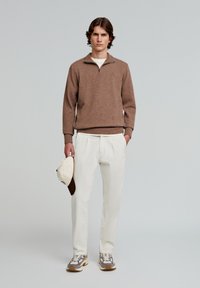 Brun sweater med lynlås, creme farvede bukser og grå-brune sneakers. Holder en lys hat med brune accenter. Enkel, ren design.