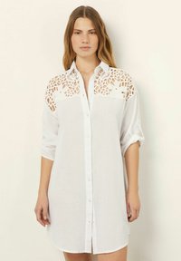 Giovane donna che indossa un vestito bianco con bottoni e maniche arrotolate, decorato con pizzo floreale sulle spalle e parte superiore del petto.