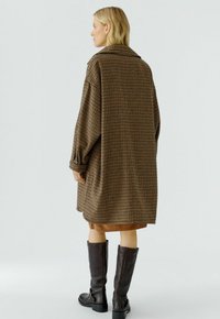 Braun karierter oversized Mantel mit Kragen, voluminösen Ärmeln und strukturiertem Stoff, kombiniert mit knielangen schwarzen Lederstiefeln.