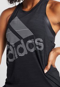 Czarny sportowy top bez rękawów z ukośnym białym logo w paski. Materiał wydaje się lekki, o gładkiej teksturze.