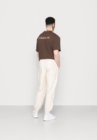 Camiseta marrón de canalé con el logotipo de "adidas" en la parte posterior, combinada con pantalones joggers crema con puños elásticos y costuras laterales, llevados con zapatillas blancas.