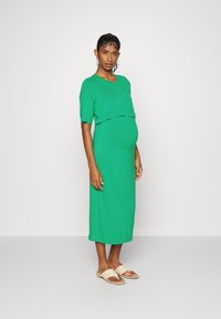 Vestido de maternidade verde com um corte relaxado e mangas curtas, apresenta um design em camadas. O tecido é suave e elástico. Combinado com sandálias bejes.