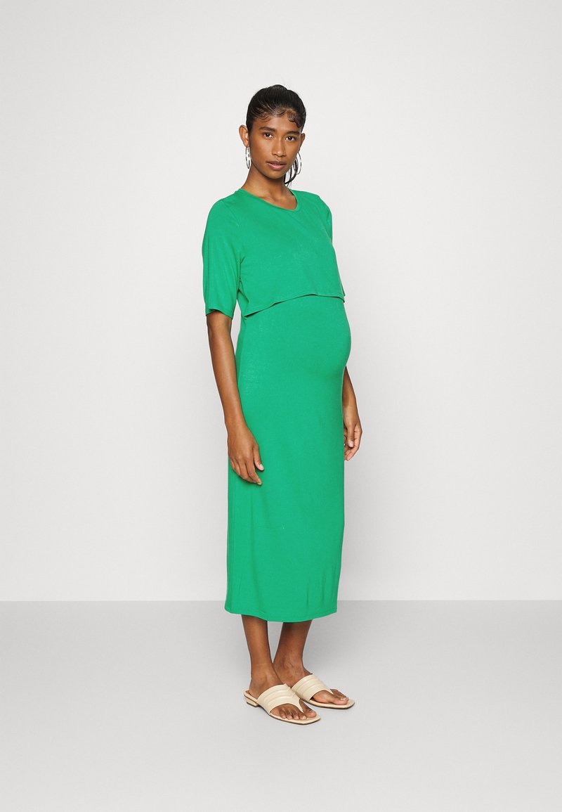 Vestido de maternidade verde com um corte relaxado e mangas curtas, apresenta um design em camadas. O tecido é suave e elástico. Combinado com sandálias bejes.