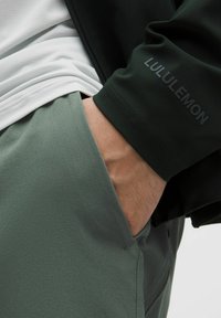 Ruka v vrecku zelených nohavíc, na sebe má čiernu bundu s logom "Lululemon" na rukáve a pod ňou bielu košeľu.