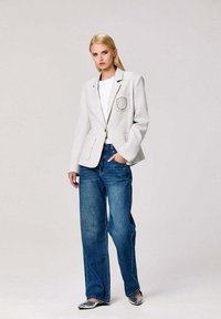 Une femme se tient debout vêtue d'un blazer blanc avec un blason, d'un haut blanc, d'un jean à jambes larges bleu, et de chaussures argentées à bouts pointus, sur un fond clair uni.