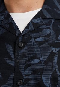 Camisa de manga corta en azul oscuro con un patrón de hojas, que presenta un collar, dos botones y una tela texturizada. Debajo lleva una camisa blanca.