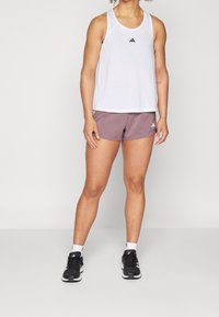 Osoba v bílém bezrukávovém topu a mauvových šortkách s černými běžeckými botami na jednobarevném pozadí.